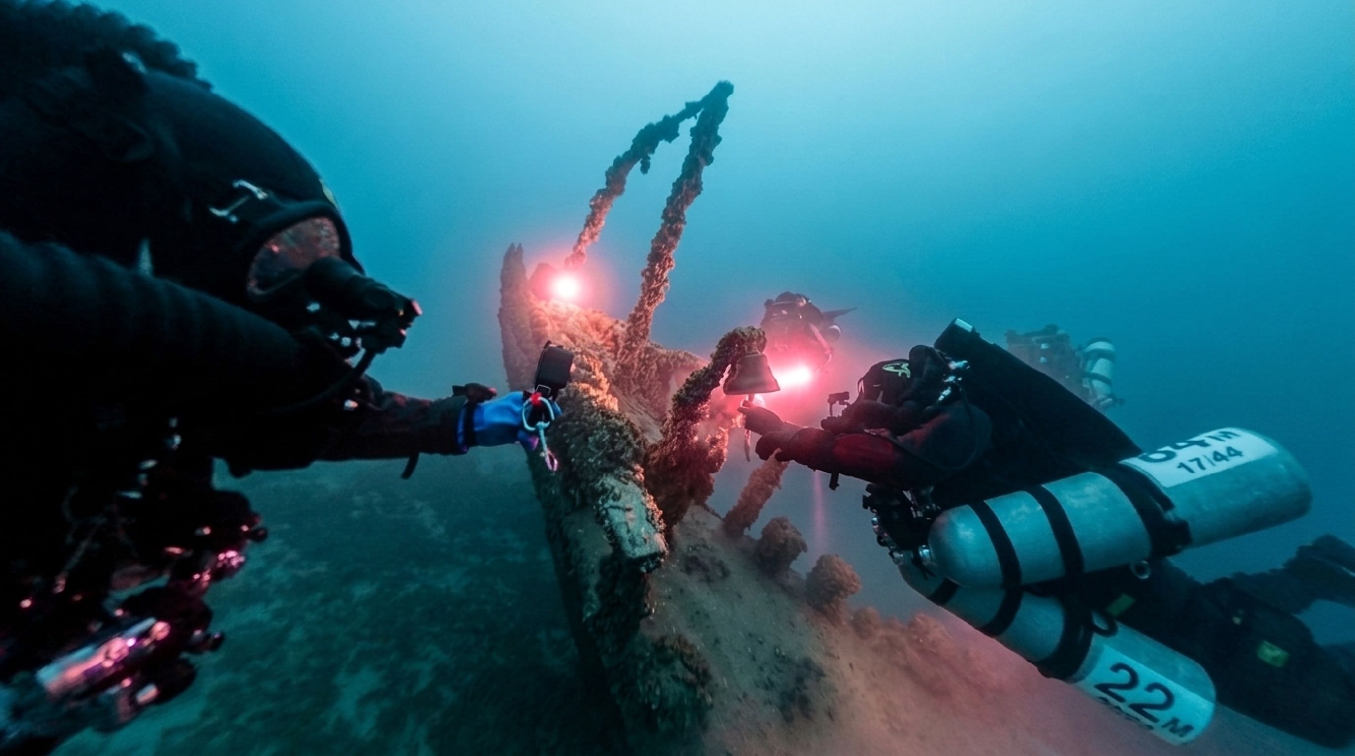 Rebreather divers explore the wreck of the Hirondelle in Lake Geneva, their red torches cutting through dark water.