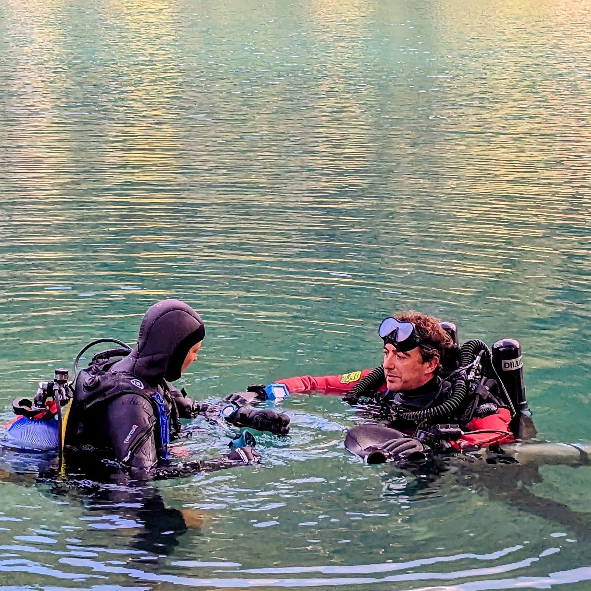 Nicholas, wearing a rebreather and a red drysuit, with a student at the surface of a lake.
